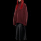 A model with long red hair and black sunglasses wears an oversized textured burgundy turtleneck sweater, a black satin midi skirt, and black pointed boots against a solid black background.