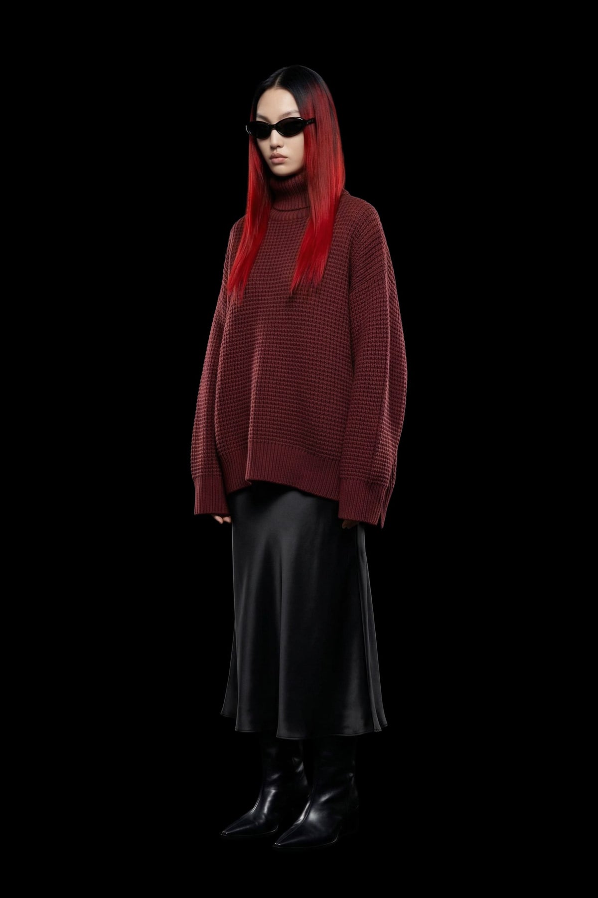A model with long red hair and black sunglasses wears an oversized textured burgundy turtleneck sweater, a black satin midi skirt, and black pointed boots against a solid black background.