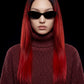 A model with long, straight black hair featuring vibrant red tips, wearing black cat-eye sunglasses and a textured dark red ribbed turtleneck sweater against a solid black background.