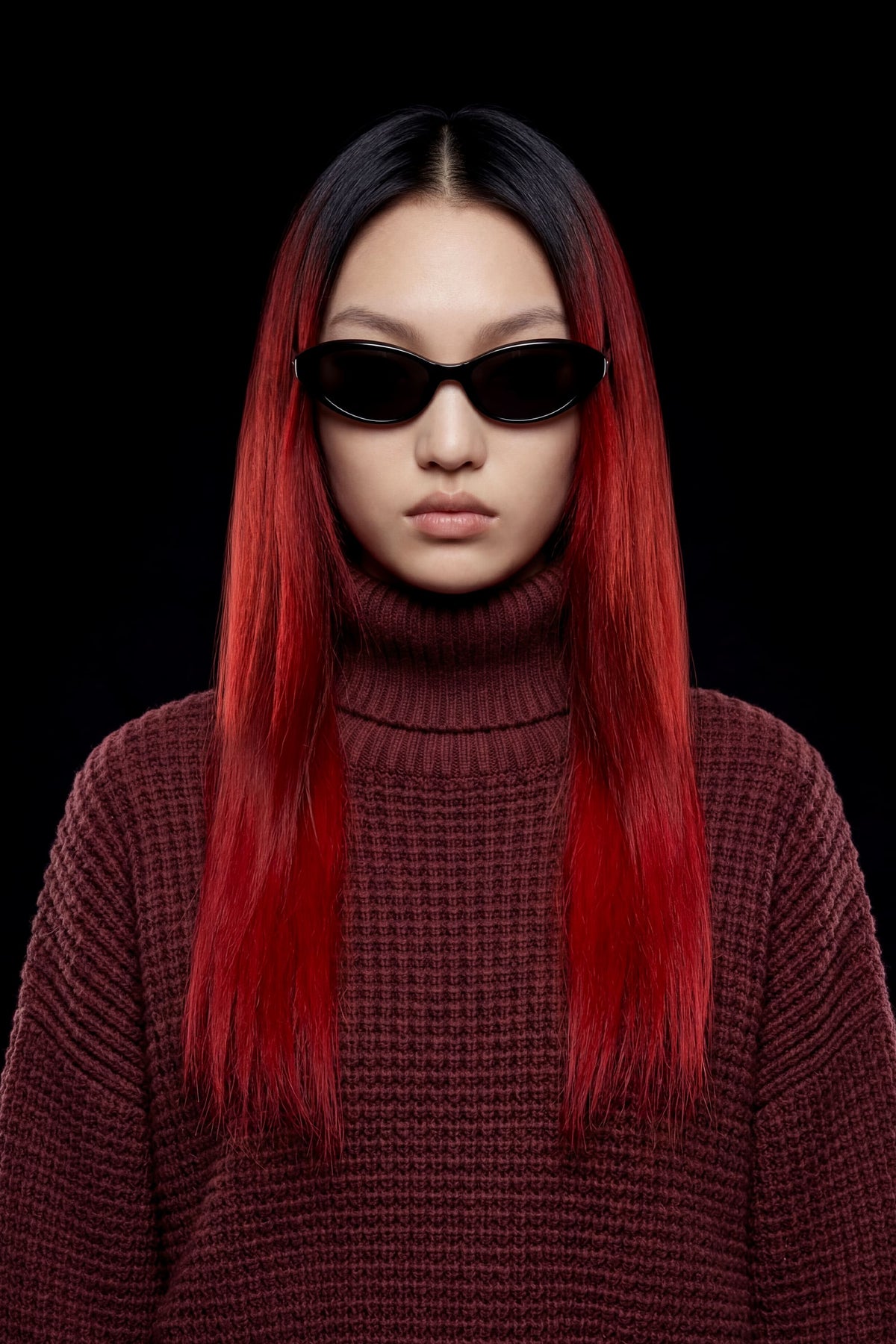 A model with long, straight black hair featuring vibrant red tips, wearing black cat-eye sunglasses and a textured dark red ribbed turtleneck sweater against a solid black background.