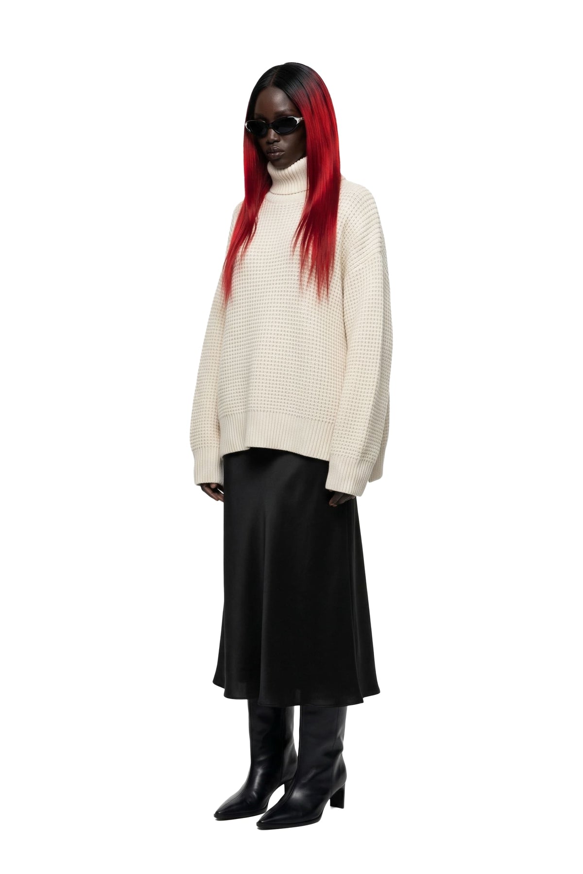 A model with long red hair wearing a cream-colored textured knit turtleneck sweater, a black midi skirt, and black pointed-toe boots, standing against a solid white background.