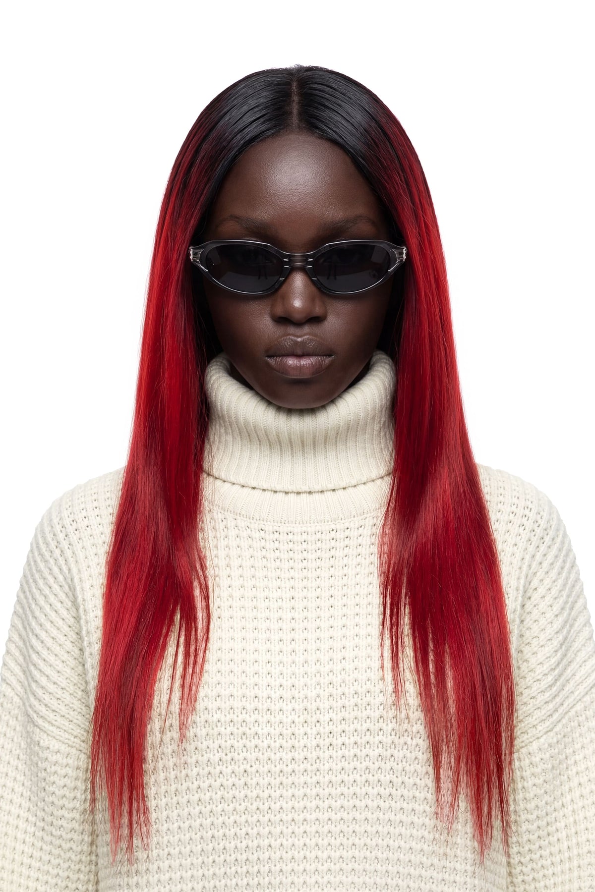 A model with long, straight black hair featuring vibrant red ombre ends, wearing a cream-colored ribbed turtleneck sweater and black sunglasses, set against a plain white background.