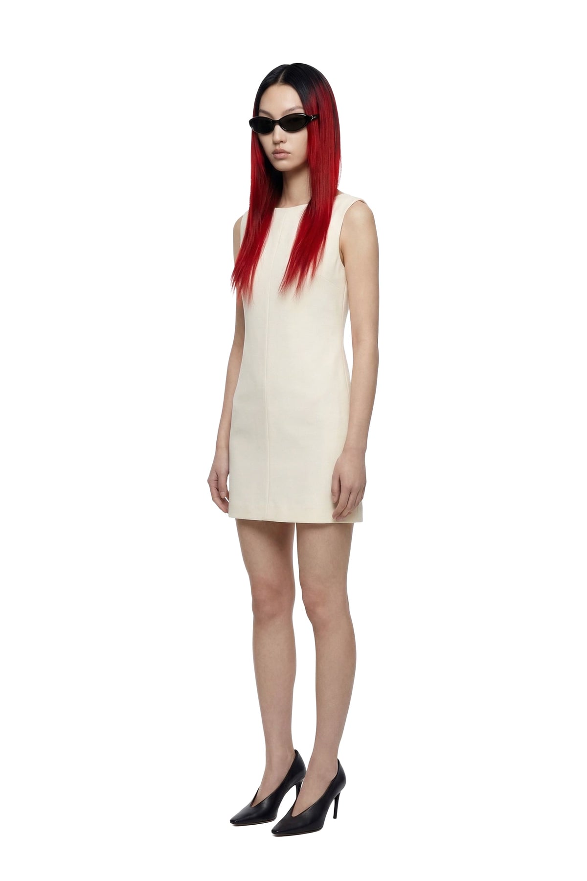 A model with long red hair and black sunglasses wearing a sleeveless cream-colored mini dress and black heels, standing against a solid white background.