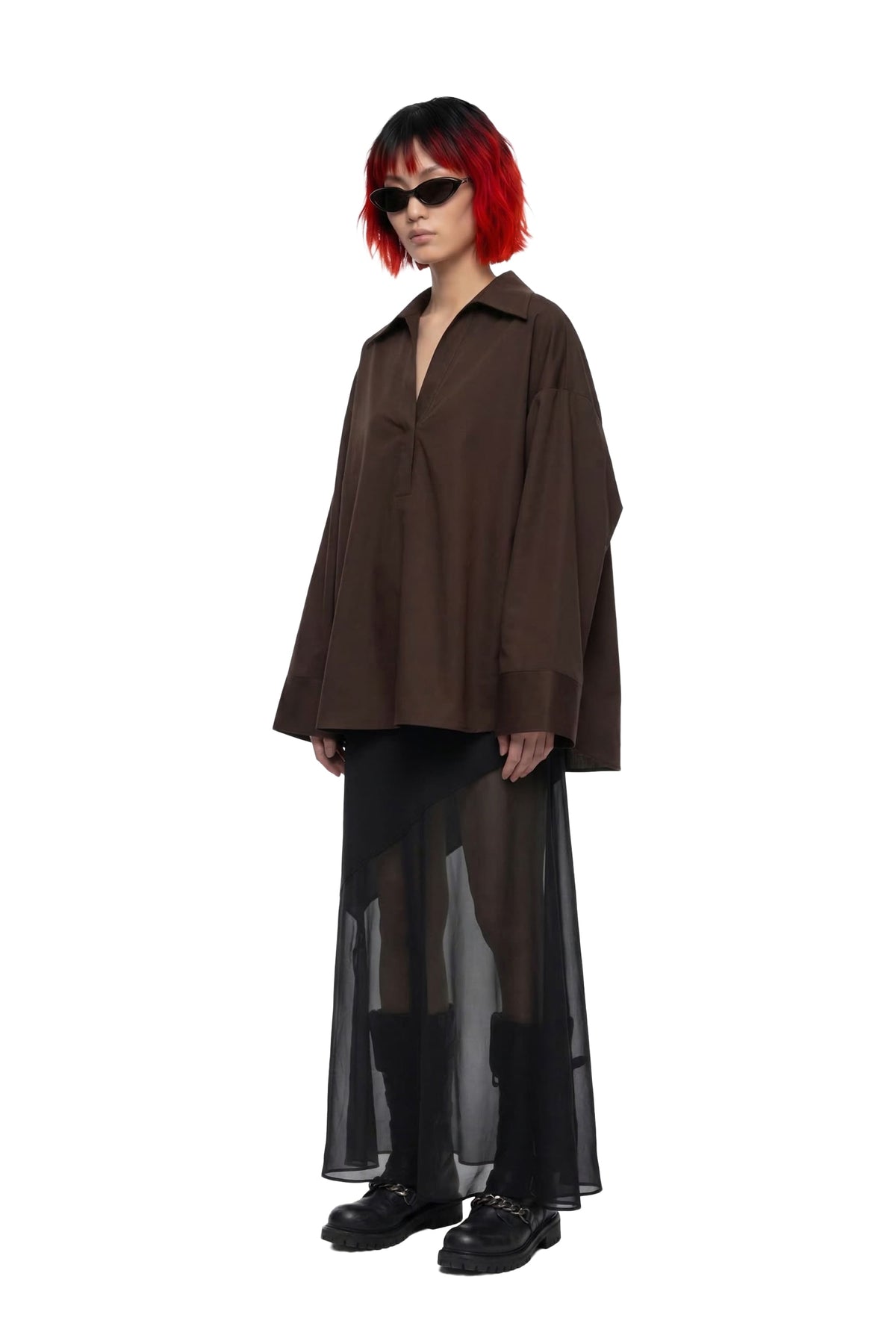A model with a red and black bob haircut wearing a dark brown oversized button-down shirt, a sheer black midi skirt, and black boots against a solid white background.
