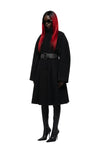 A model wearing a long black coat with a wide black belt, black knee-high boots, and black sunglasses, featuring vibrant red hair, shot against a solid white background.