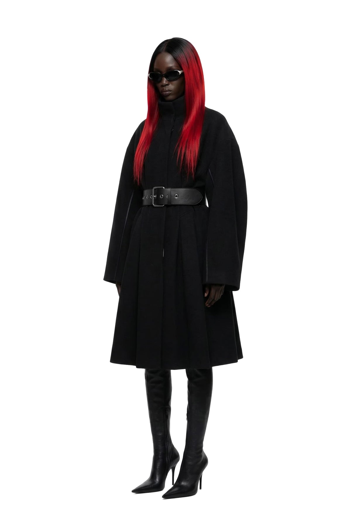 A model wearing a long black coat with a wide black belt, black knee-high boots, and black sunglasses, featuring vibrant red hair, shot against a solid white background.