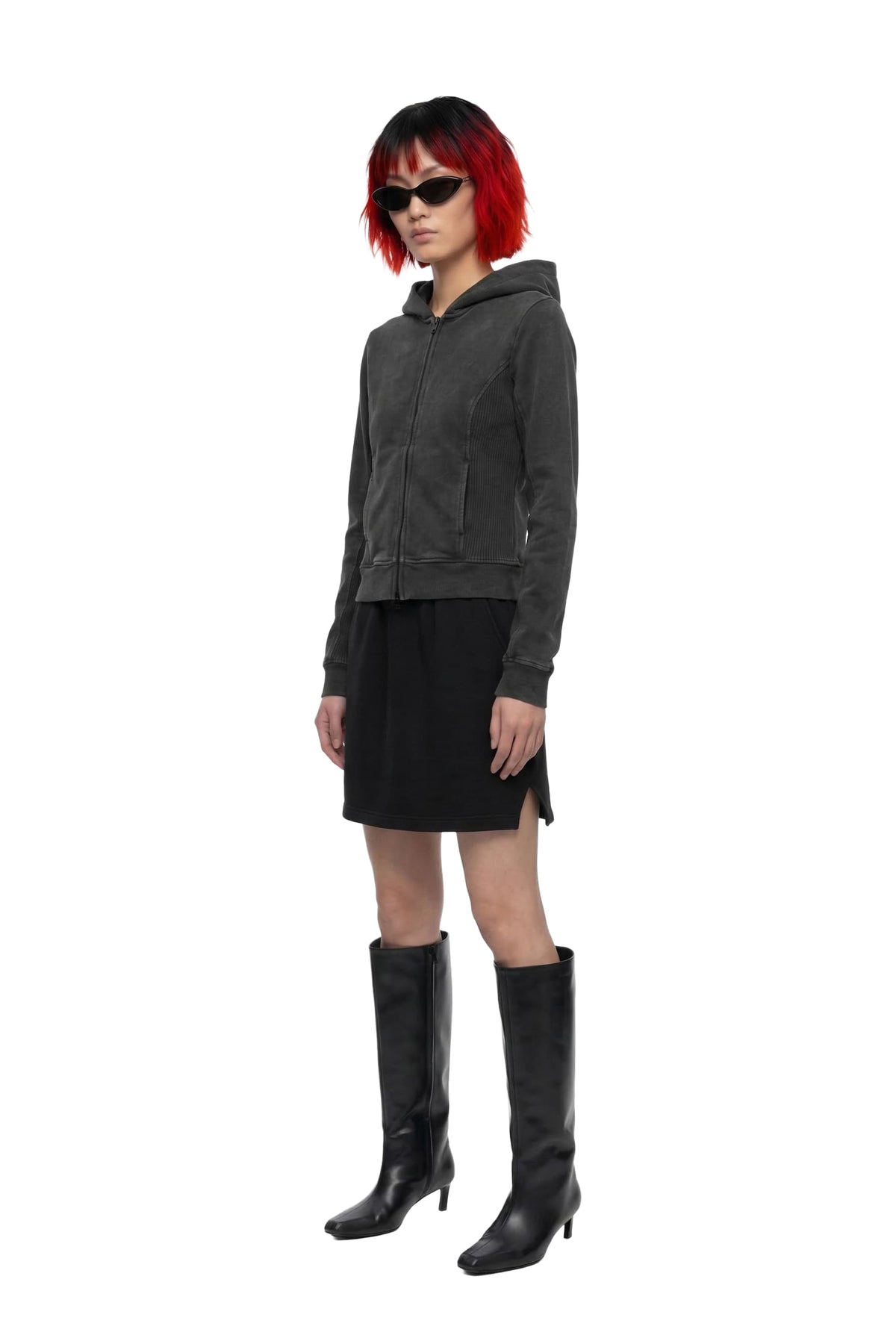 A model with short red hair and black sunglasses is shown in profile wearing a dark gray cropped hooded jacket, a black mini skirt, and black knee-high heeled boots against a plain white background.
