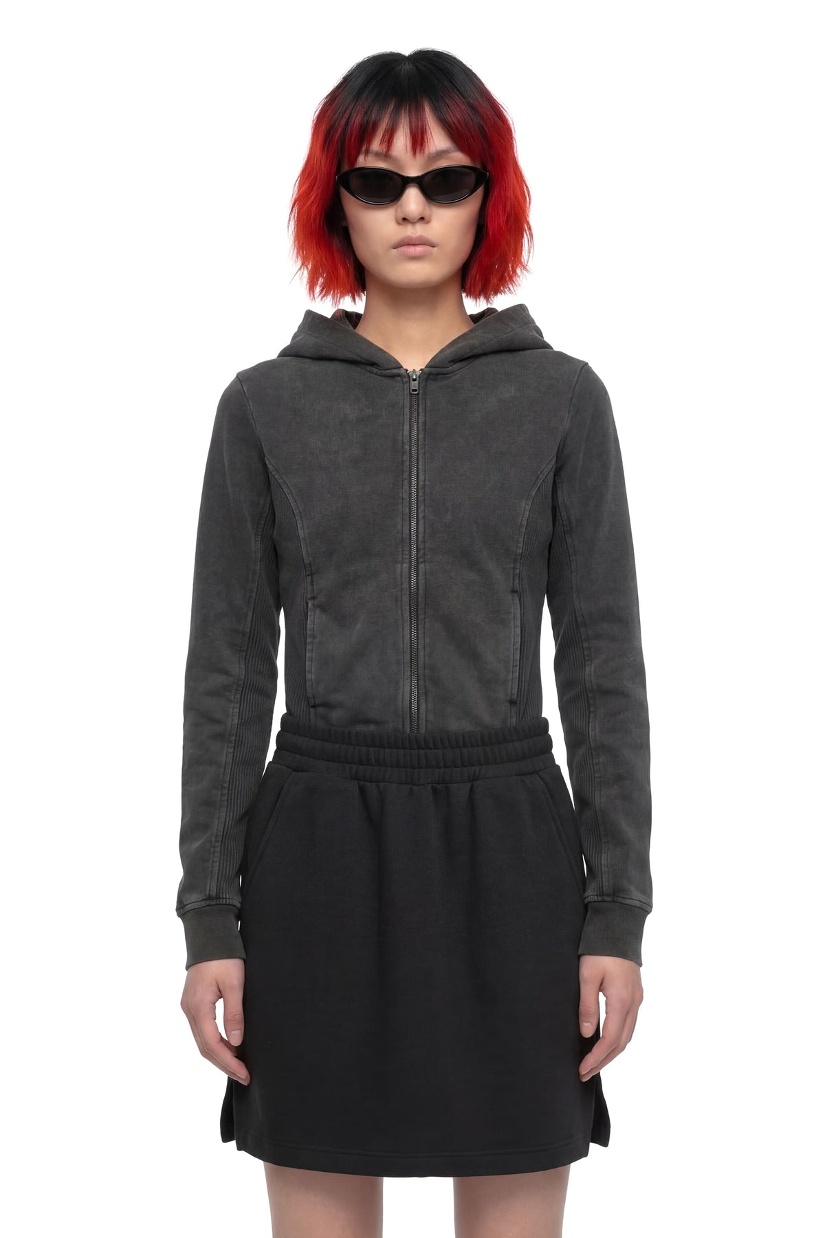 A model with red and black hair wearing a dark gray zip-up hooded sweatshirt and a black skirt, photographed against a solid white background.