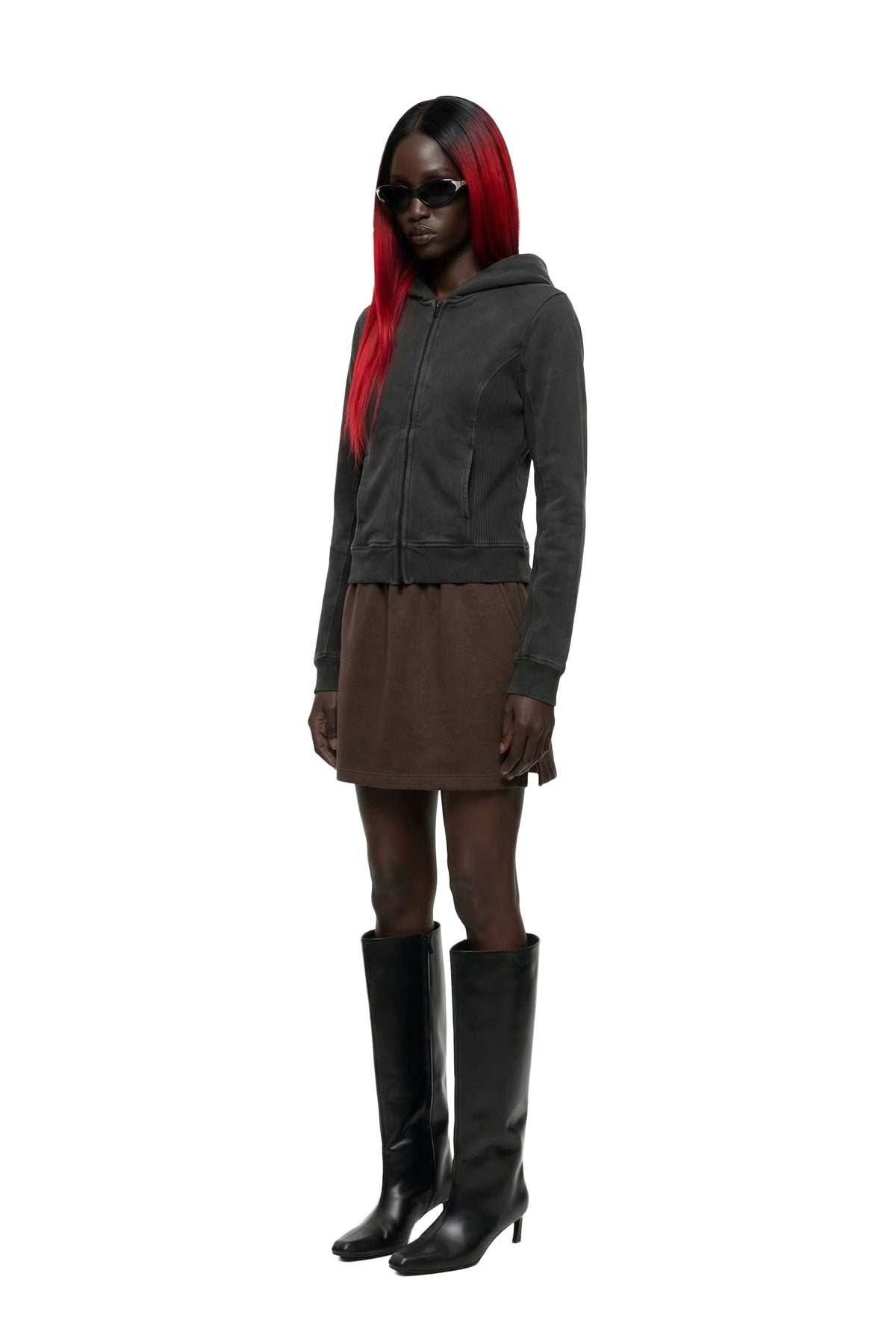 A model with long red hair and black sunglasses stands against a white background wearing a dark gray zip-up hoodie, a brown mini skirt, black tights, and black knee-high boots.