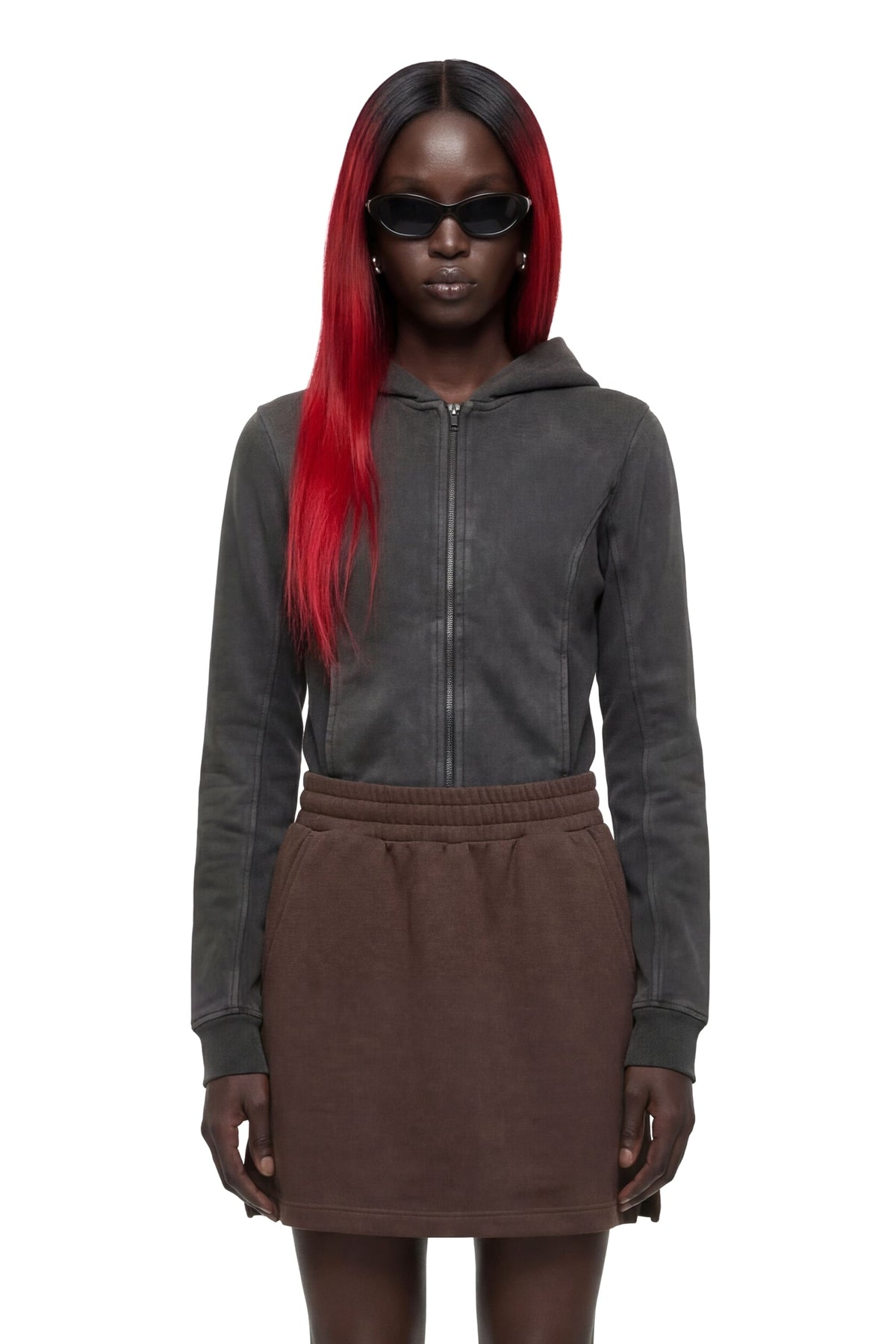A model with long red hair and black sunglasses wears a dark gray zip-up hoodie and a brown mini skirt against a plain white background.