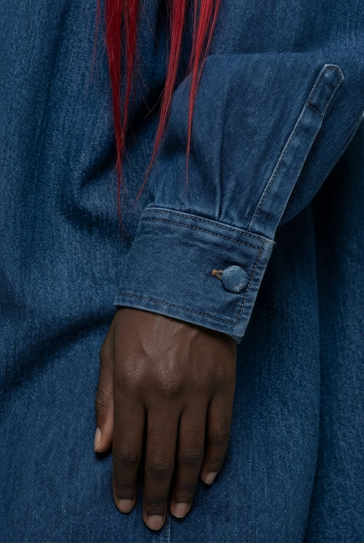 Close-up of a person wearing a blue denim shirt or jacket, showing the sleeve cuff with a button closure and a hand resting below it. Red hair is visible at the top edge.
