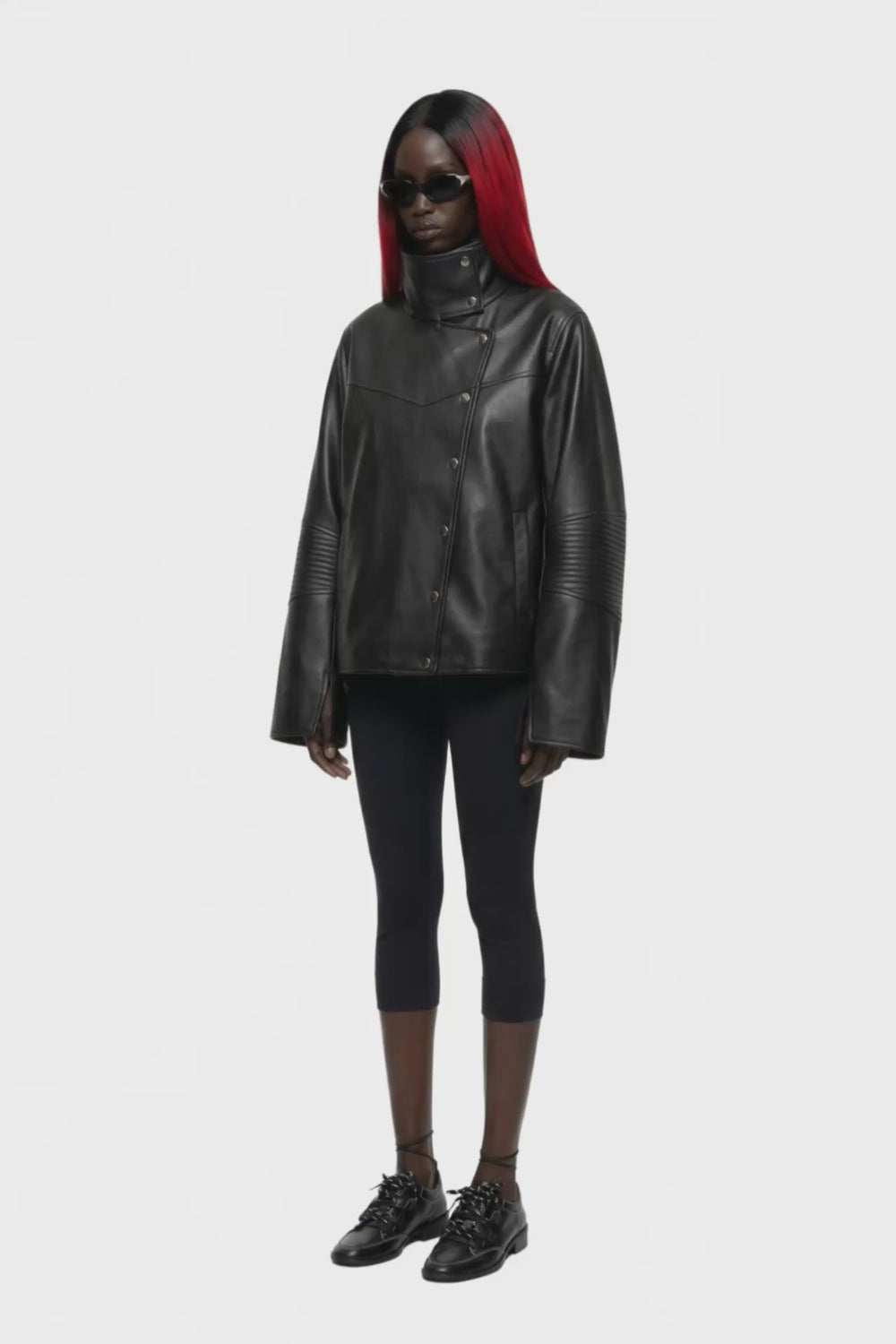 A black leather jacket with a high mock neck, asymmetrical snap button closure, and ribbed sleeve detailing, shown against a plain white background.