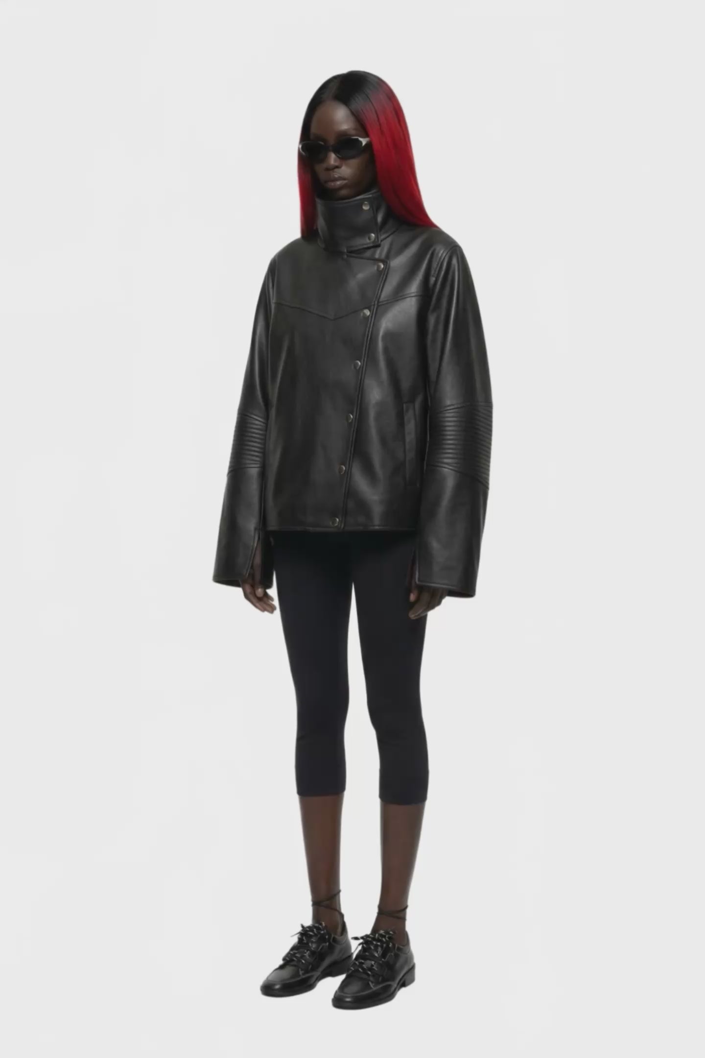 A black leather jacket with a high mock neck, asymmetrical snap button closure, and ribbed sleeve detailing, shown against a plain white background.