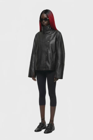 A black leather jacket with a high mock neck, asymmetrical snap button closure, and ribbed sleeve detailing, shown against a plain white background.