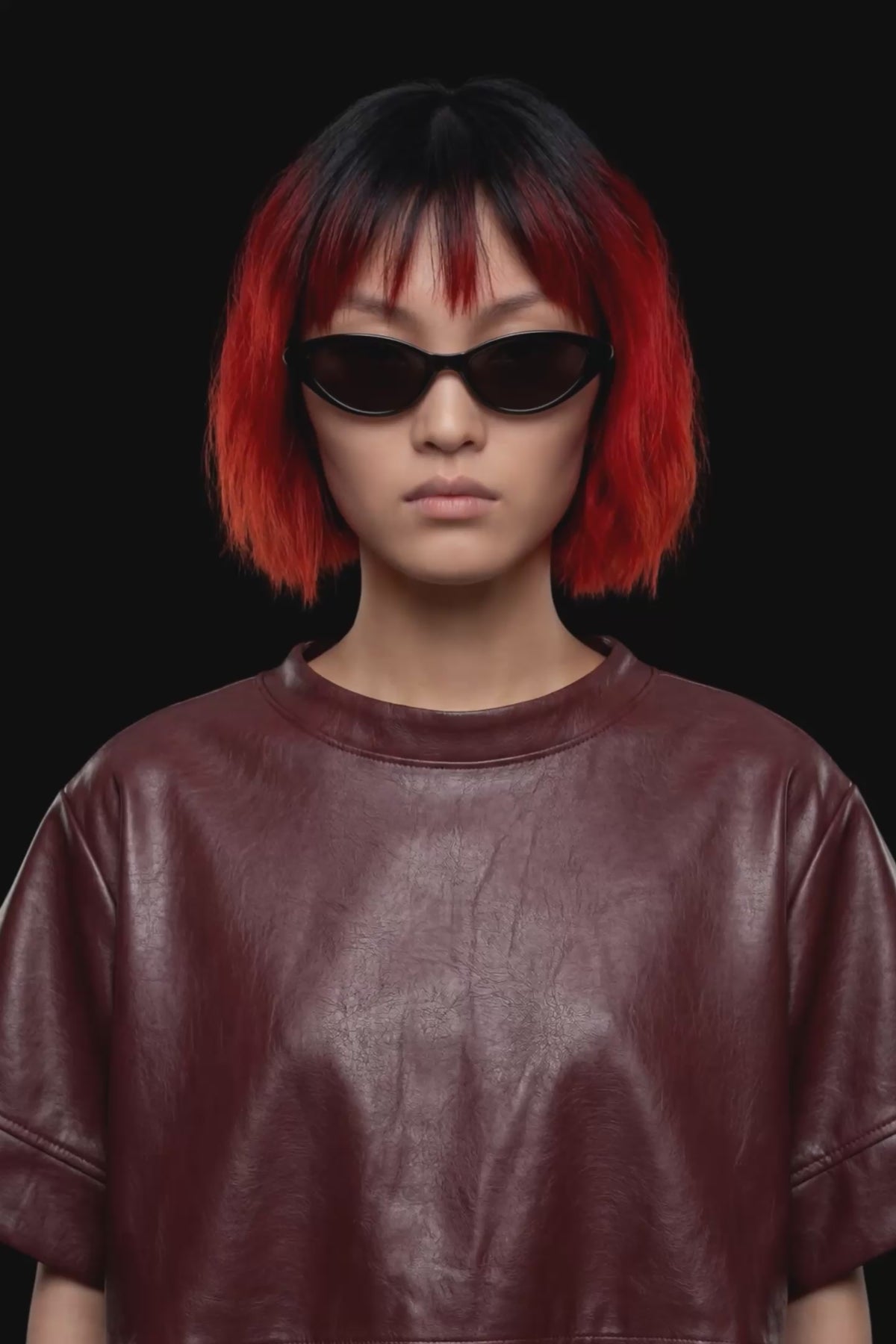 A short-sleeve cropped faux-leather top in a deep burgundy color, featuring a layered asymmetrical hem, displayed against a solid black background.