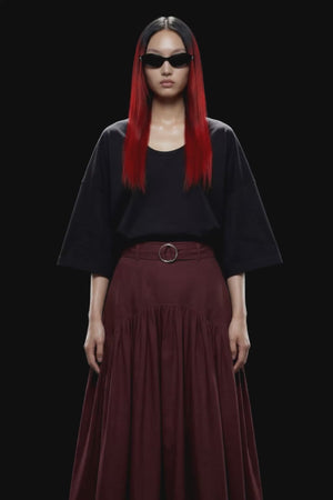 A dark burgundy pleated midi skirt with a matching waistband belt and silver-tone ring buckle, shown against a solid black background.