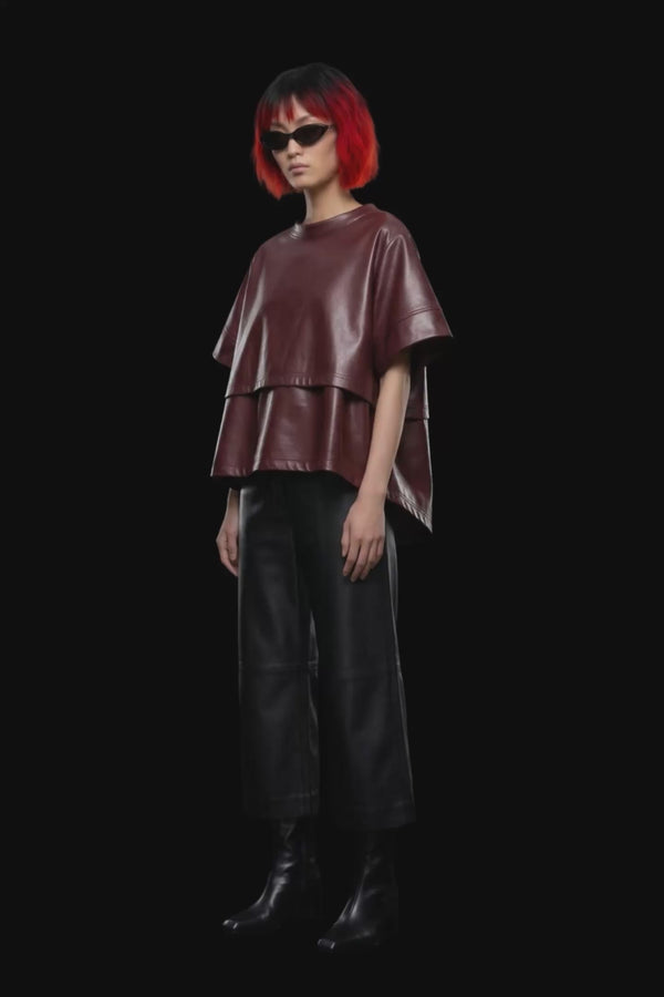 A short-sleeve cropped faux-leather top in a deep burgundy color, featuring a layered asymmetrical hem, displayed against a solid black background.