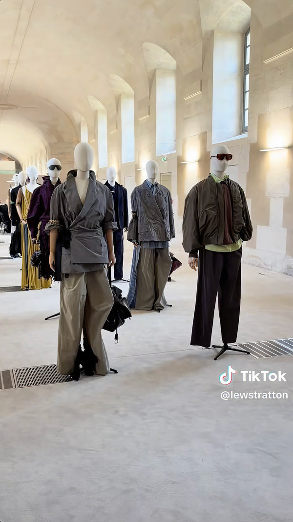 A fashion runway scene in a large arched hall with mannequins wearing layered clothing. The most prominent outfit is a dark brown jacket over a light green shirt with dark wide-leg trousers, while other mannequins wear gray and beige ensembles.