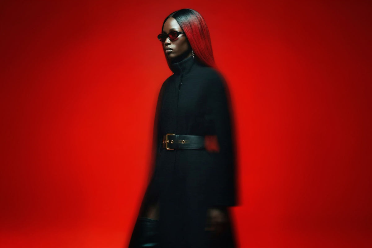 A model walks through a minimalist red studio setting wearing a long black coat with a wide silver belt, black leather pants, and black boots.
